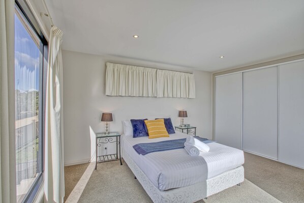 A bedroom with a large bed, two bedside tables with lamps, a window with curtains, and a wardrobe. - Georges Bay Luxury - St Helens (Break O'Day)
