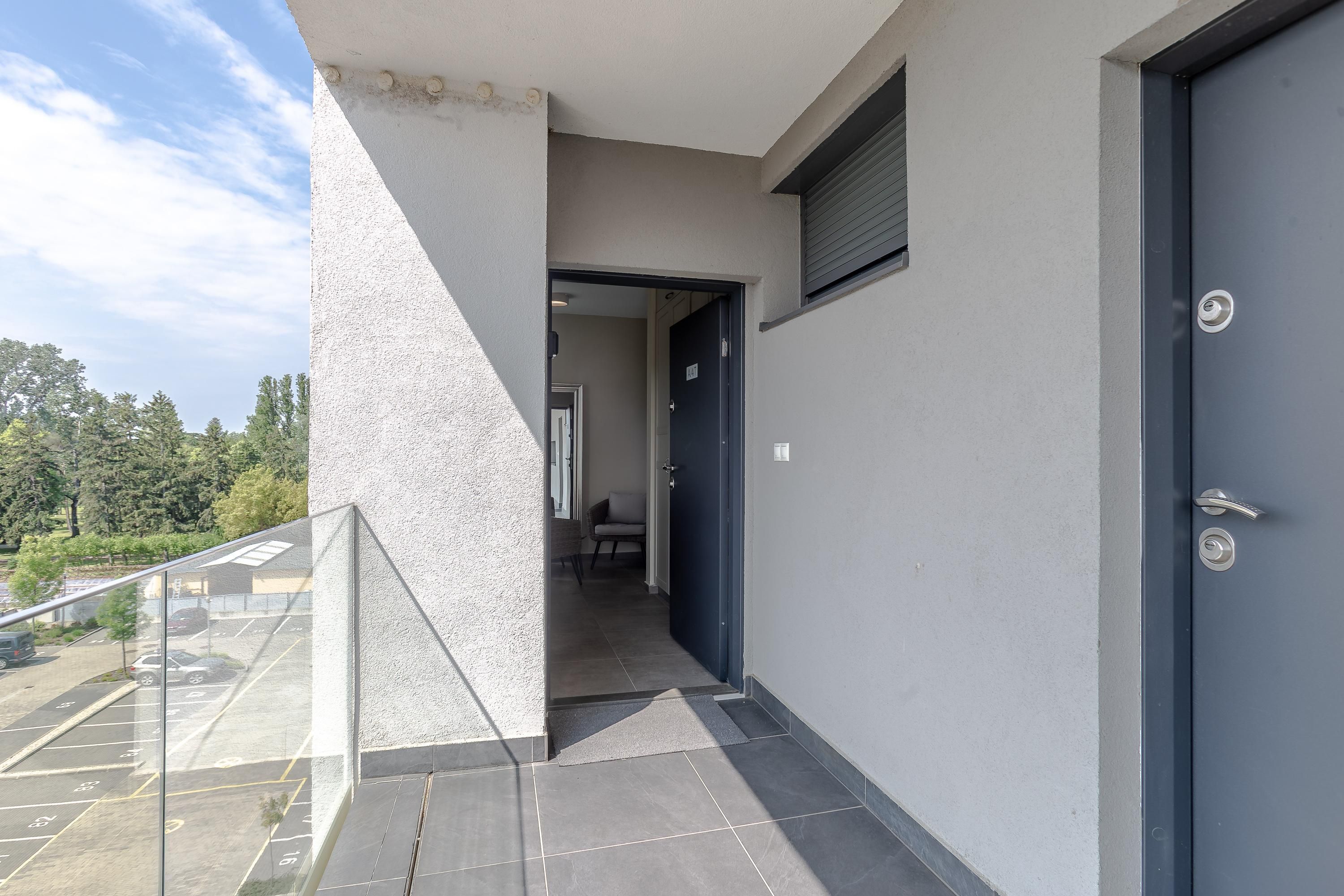 Fachada de un edificio moderno con una puerta azul oscuro, un balcón de cristal y un estacionamiento visible a través de las ventanas.