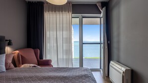 A hotel room with a bed, a chair, a radiator, and a view of the sea through a large window. - Lakefront Elegant Condo Lela (Balatonlelle)