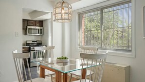Un área de comedor con una mesa de vidrio y sillas, una lámpara colgante, una ventana con persianas y una cocina al fondo.