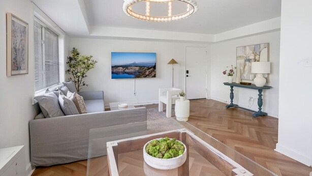 Una sala moderna con un sofá, una mesa de centro de vidrio con un cuenco, una araña, un cuadro y una consola decorada con objetos ornamentales.