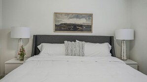 Una cama bien hecha con sábanas blancas, cabecera gris y almohada estampada. Hay dos mesitas de noche con lámparas y un jarrón con flores. Sobre la cama cuelga un cuadro enmarcado con un paisaje.
