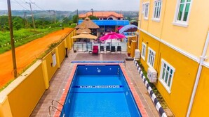 A swimming pool with a blue interior, surrounded by a yellow building with a thatched roof structure and outdoor seating area. - Remo Majestic Hotel (Sagamu)