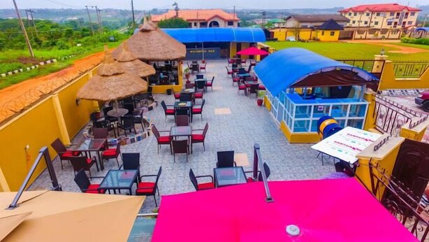 An outdoor dining area with tables and chairs, a thatched-roof structure, and a blue-roofed building in the background. - Remo Majestic Hotel (Sagamu)