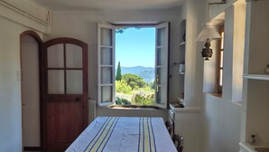 Een eethoek met een tafel gedekt voor twee personen, uitzicht op groen en bergen door een open raam, en een houten deur.