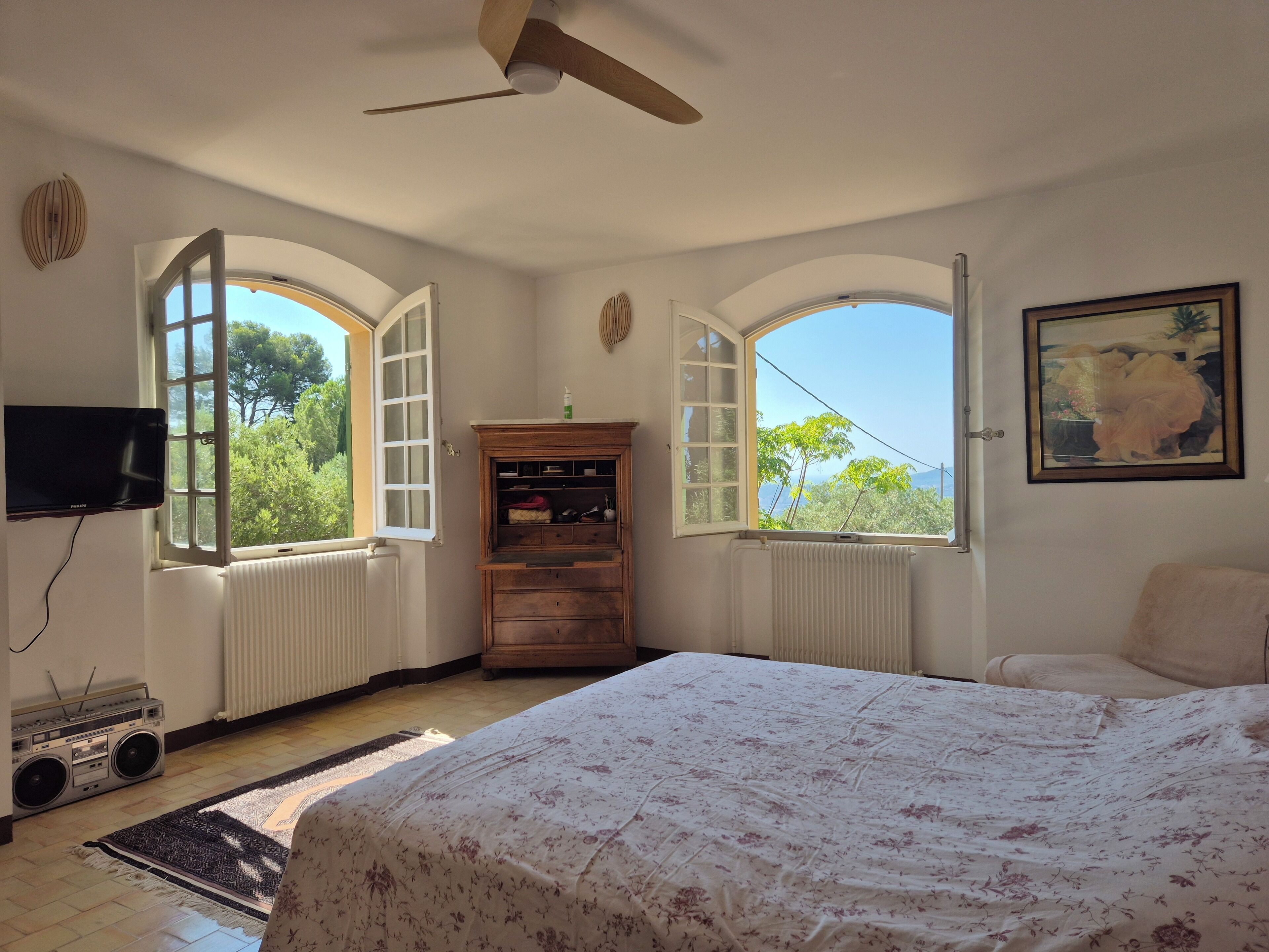 A bedroom with a bed, a wooden cabinet, a television, and two windows with arched tops.