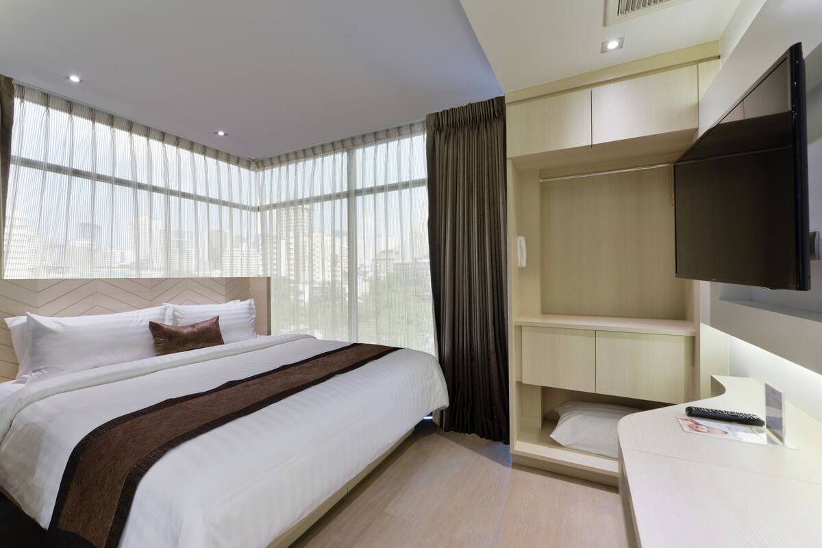 Habitación de hotel con una cama grande, televisión, escritorio y vistas a la ciudad a través de cortinas translúcidas.