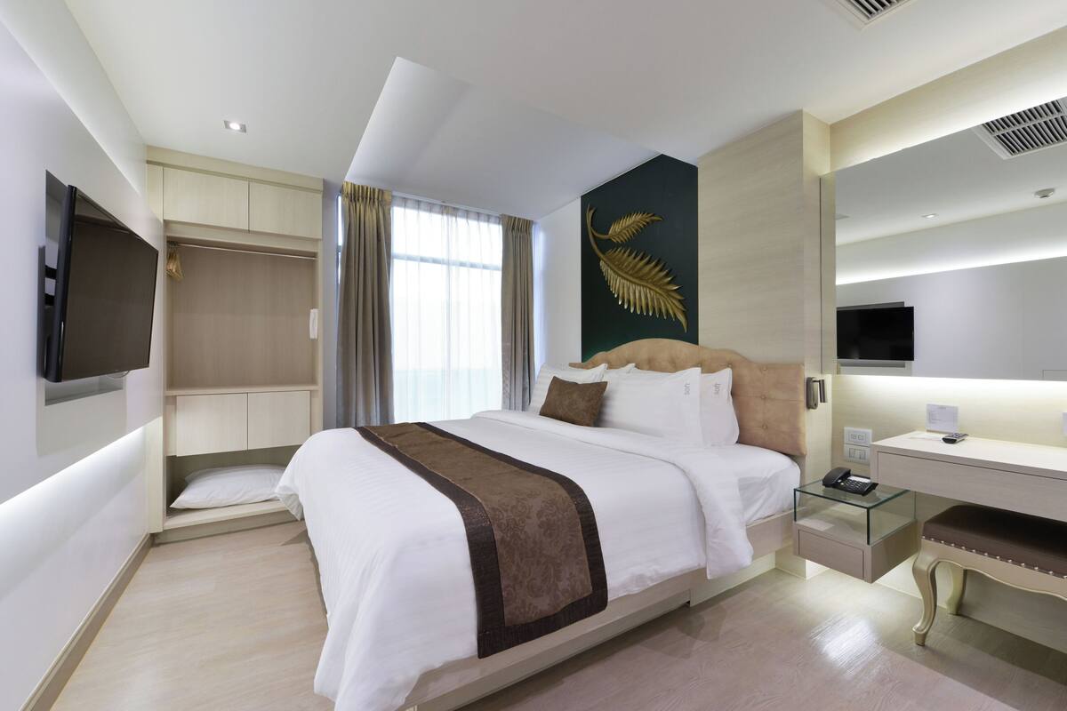 Una habitación de hotel moderna con una cama grande, un escritorio de cristal, un televisor de pantalla plana en la pared y un amplio ventanal con cortinas.