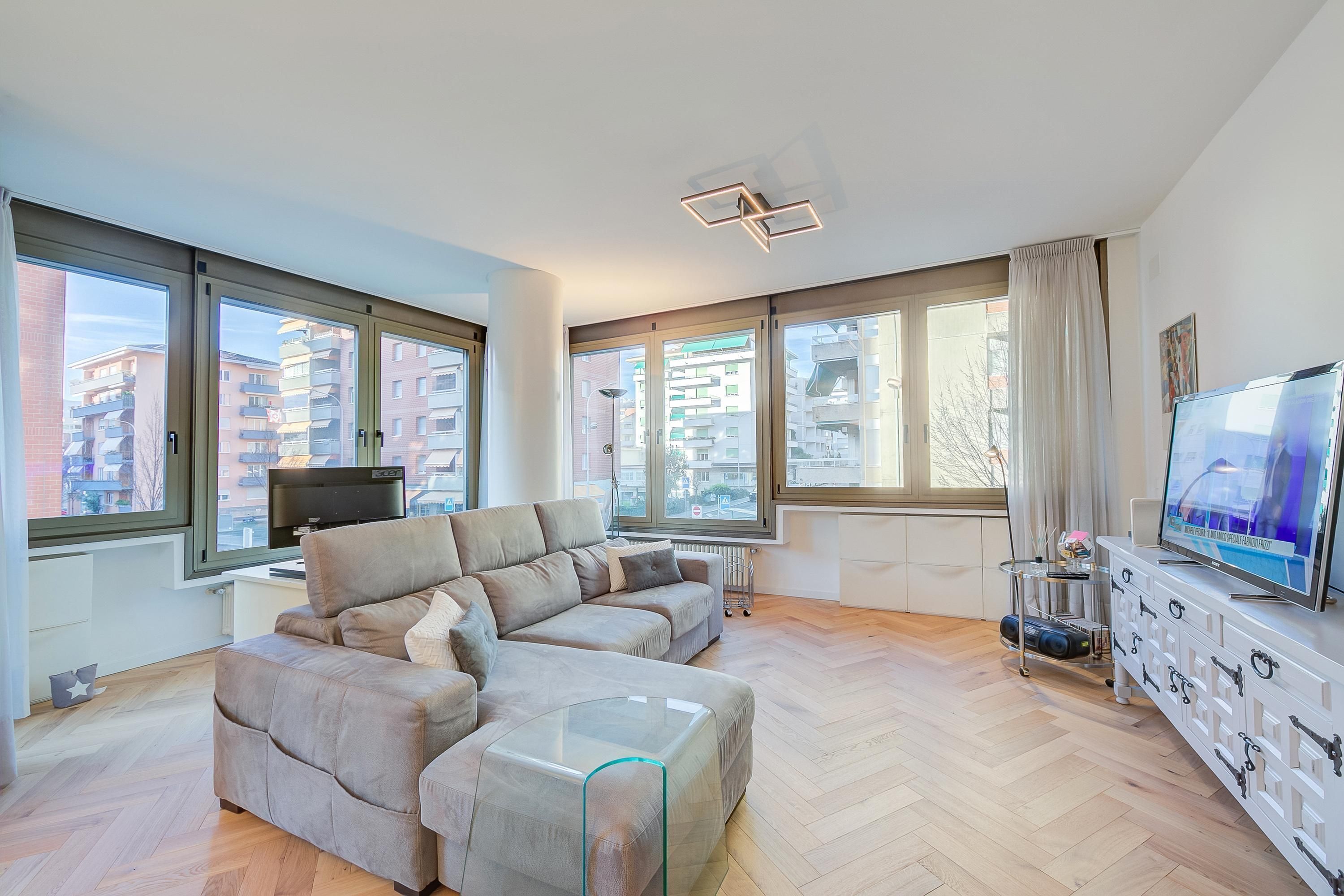 A modern living room with a sectional sofa, a glass coffee table, and a TV on a white cabinet. Large windows offer a view of the city.