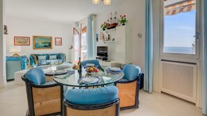 A dining area with a glass table, blue upholstered chairs, and a chandelier. There is a sofa and a television in the background. - Rhapsody By The Sea in Sanremo (Sanremo)