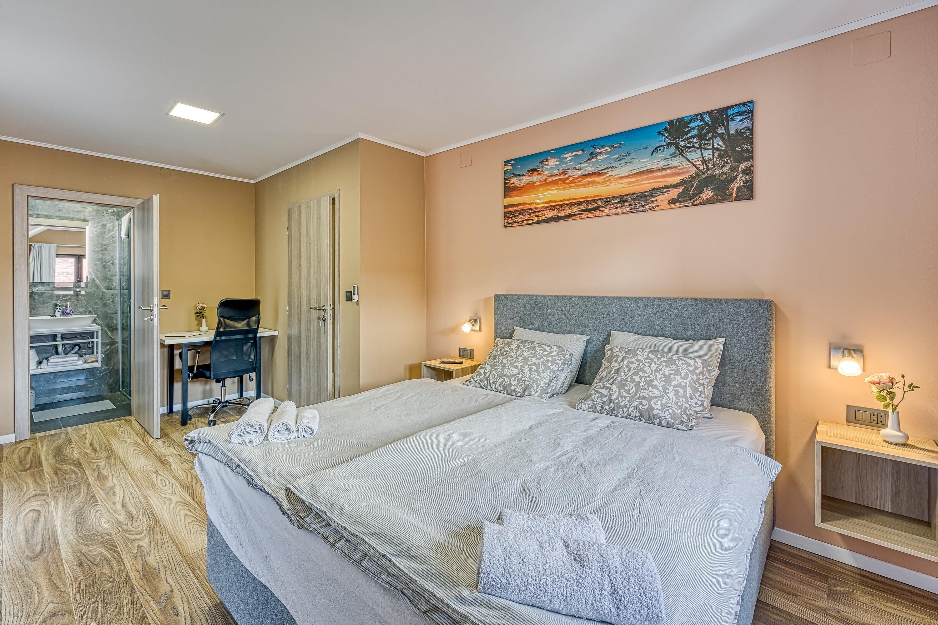 A bedroom with a large bed, a desk, and a chair, a painting on the wall, and a door leading to a bathroom.