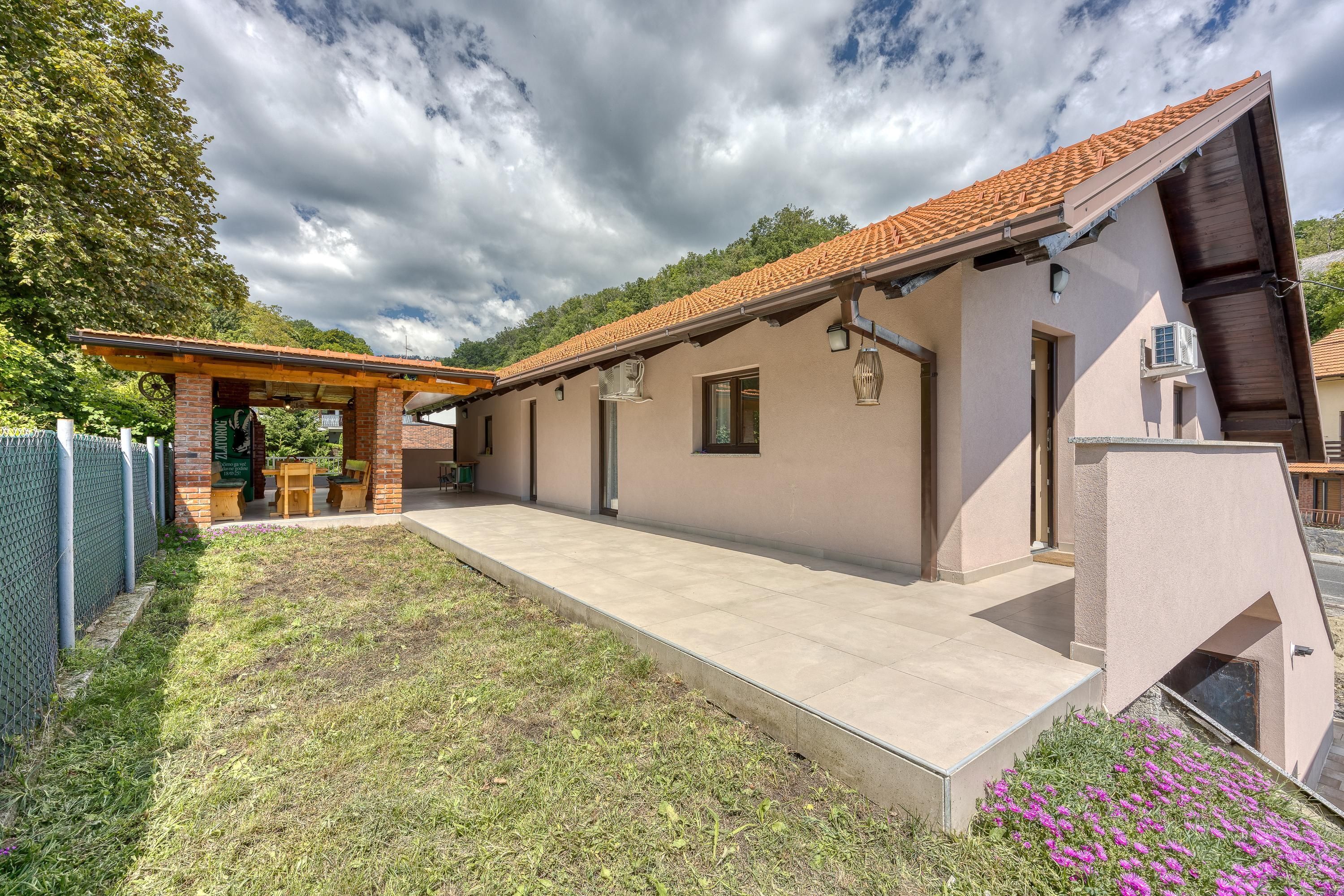 Ein modernes Haus mit überdachter Terrasse, einem eingezäunten Garten und einem kleinen Garten mit violetten Blumen.