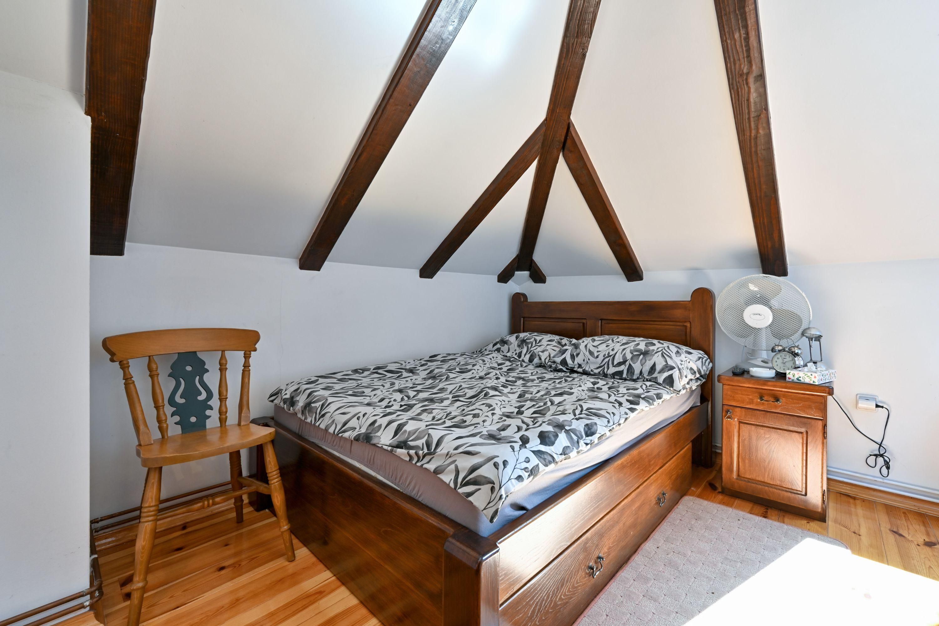 A bedroom with a wooden bed, a wooden chair, a bedside table with a fan, and a small cabinet.