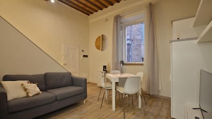 Un espacio habitable funcional con un sofá gris, una mesa de comedor con sillas, una ventana con cortinas y un mueble blanco.