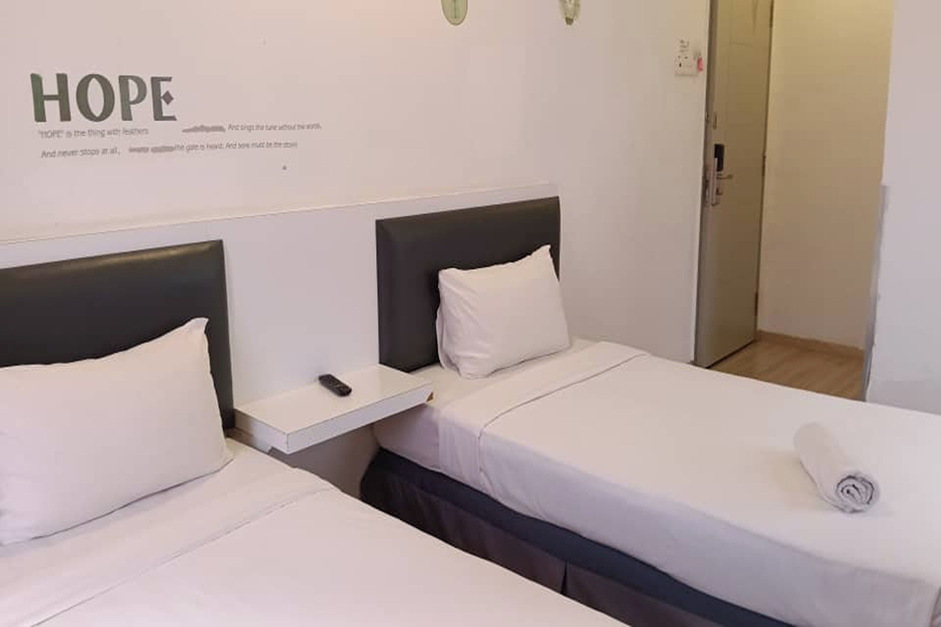 A hotel room with two single beds, a small bedside table with a remote control, and a wall with motivational text.
