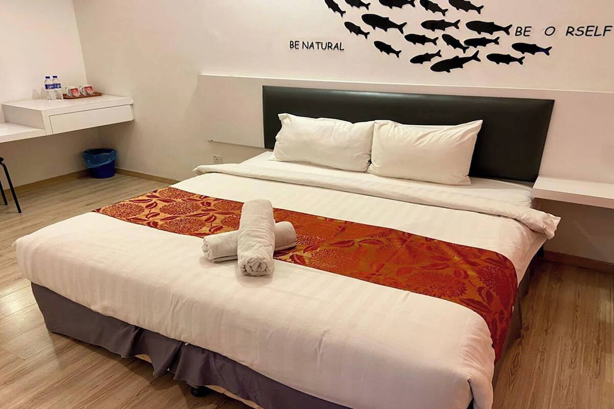 A hotel room with a bed, a headboard, a wall decoration, a small table with toiletries, and a trash bin.
