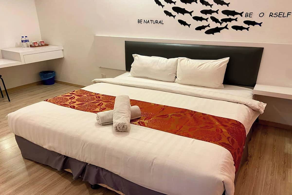 A hotel room with a bed, a headboard, a wall decoration, a small table with toiletries, and a trash bin.