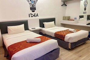 Ein Hotelzimmer mit zwei Betten, jeweils mit weißer Bettwäsche und einem gemusterten Überwurf. Über den Betten befindet sich eine Wandleuchte mit dem Schriftzug "IDEA" und ein Logo. Ein kleiner Tisch mit Telefon und Tasse steht zwischen den Betten.