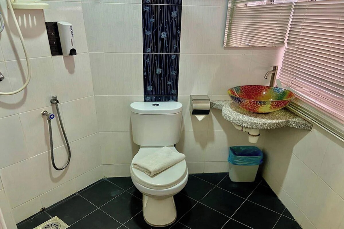 Une salle de bain comprenant des toilettes, une douche, un lavabo avec un récipient décoré de mosaïques, une poubelle et un sèche-mains.