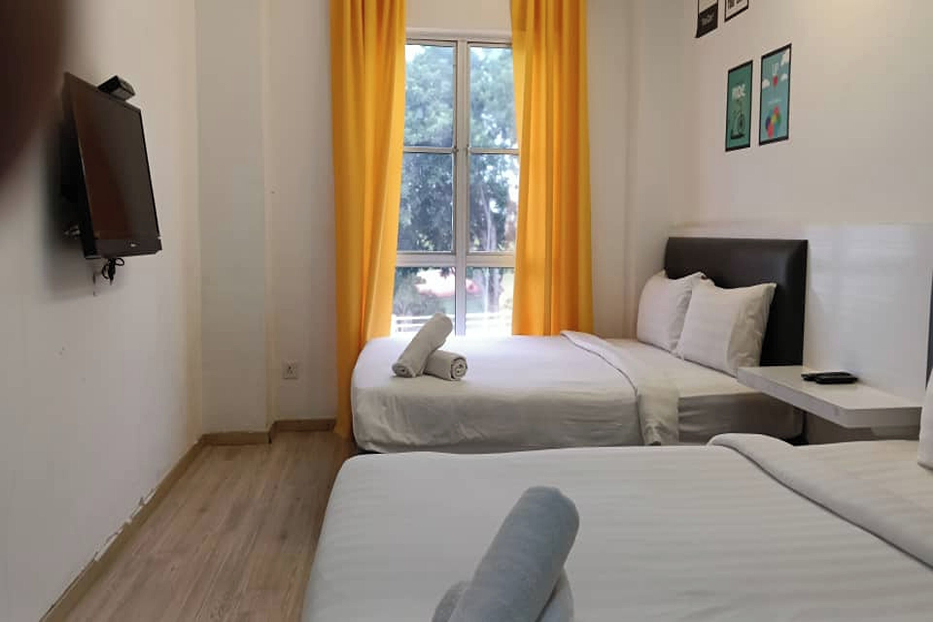 A hotel room with two beds, a window with yellow curtains, a flat-screen TV mounted on the wall, and a small table with a remote control.