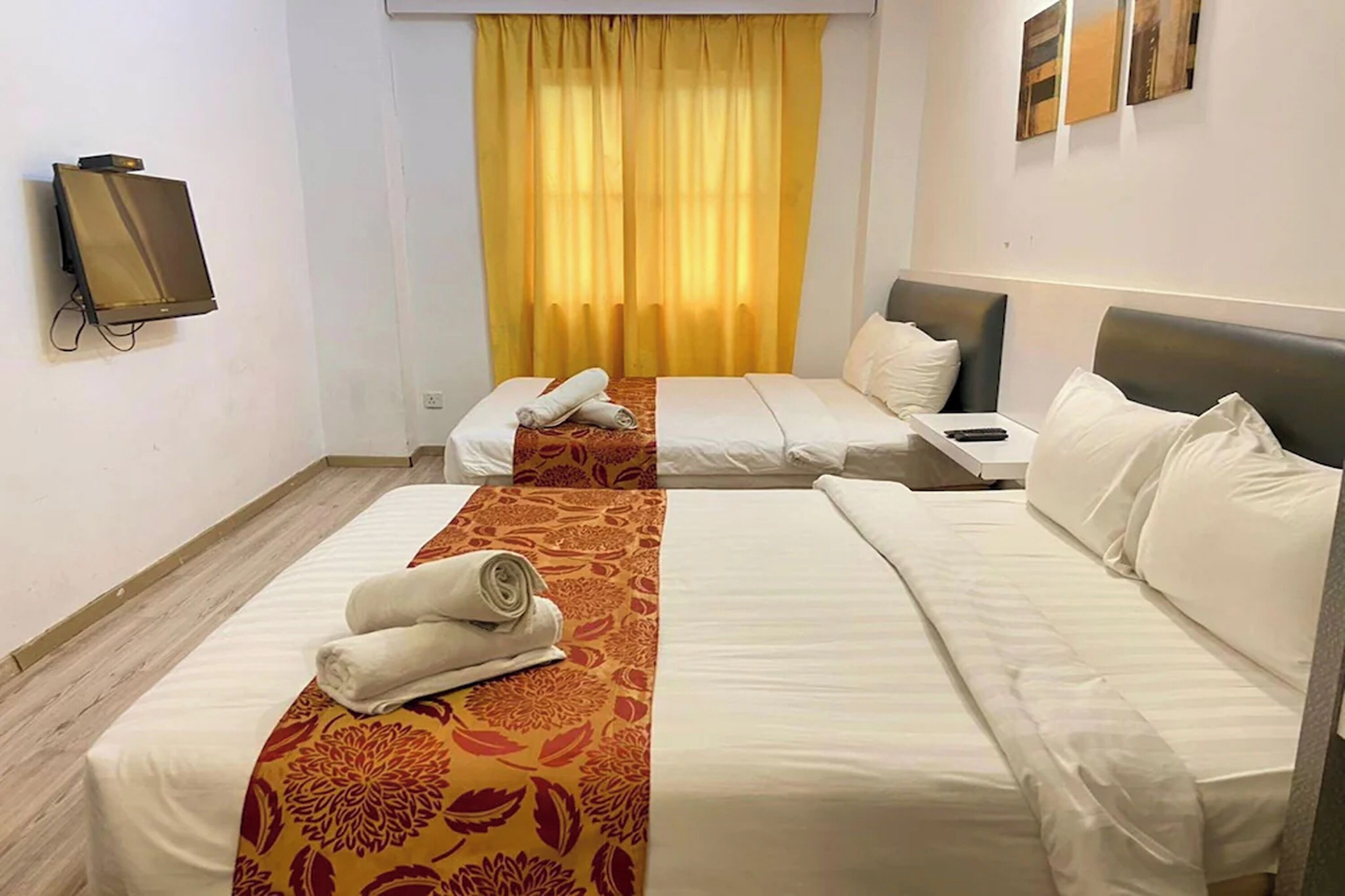 A hotel room with two beds, a TV mounted on the wall, and a window with yellow curtains.
