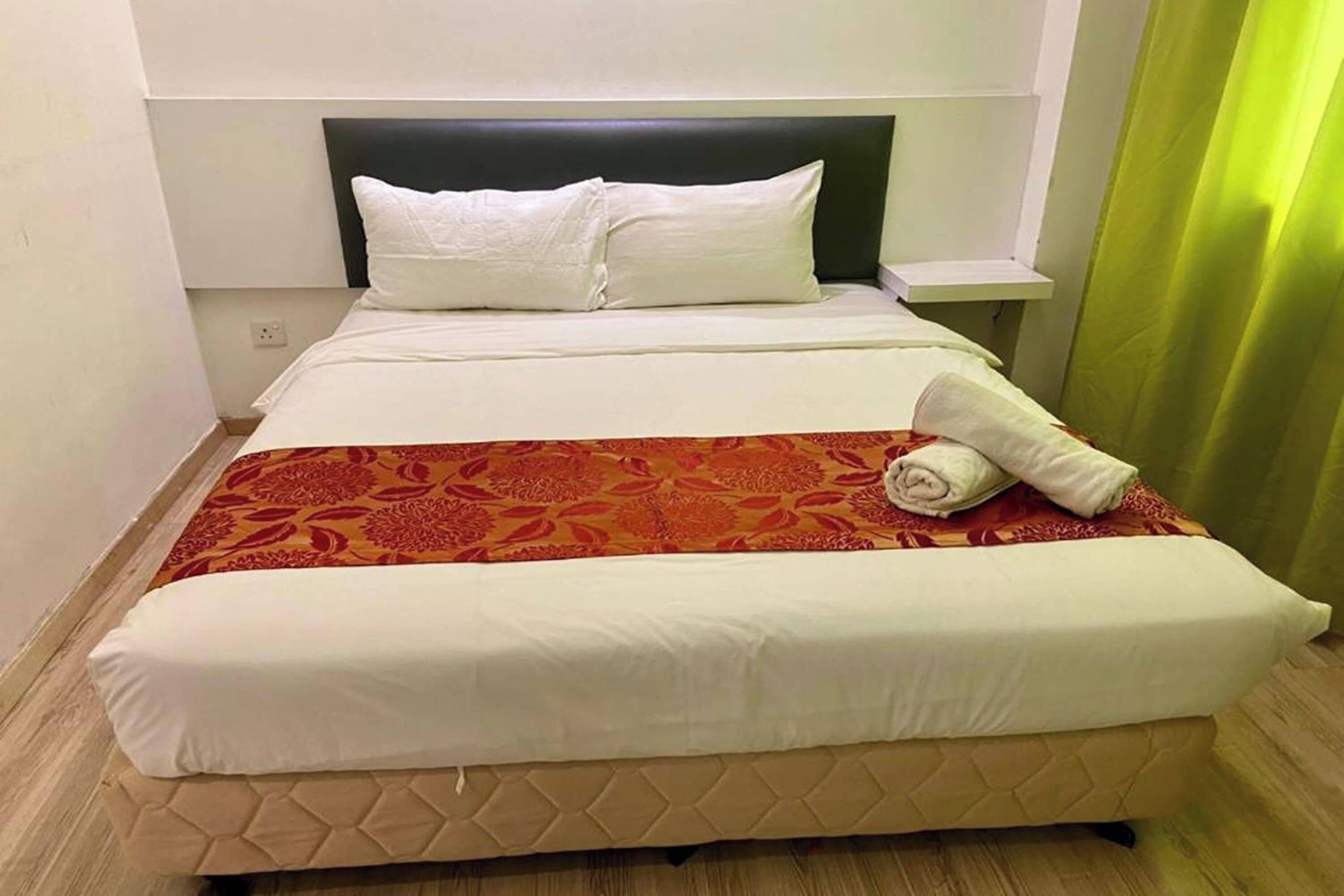 A neatly made bed with white linens, a red patterned bedspread, and a rolled towel on the side.