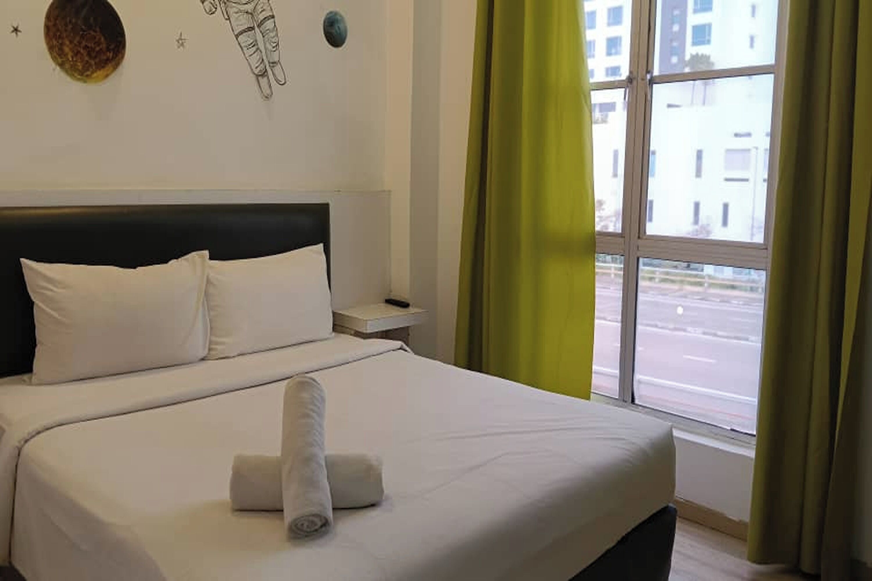 Habitación de hotel con cama, ropa de cama blanca, mesita de noche, reloj de pared y ventana con cortinas.