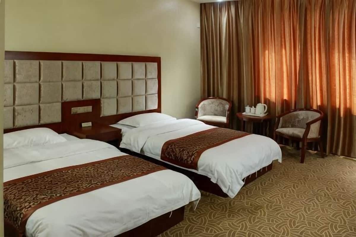 A hotel room with two beds, a wooden headboard, a small table with a chair, and a window with curtains.