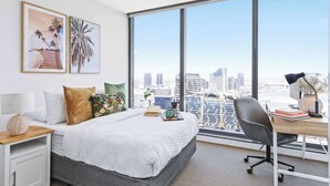 Ein modernes Schlafzimmer mit einem großen Bett, einem Schreibtisch und Blick auf eine Stadtlandschaft durch große Fenster.