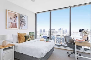 A modern bedroom with a large bed, a desk, and a view of a cityscape through large windows.