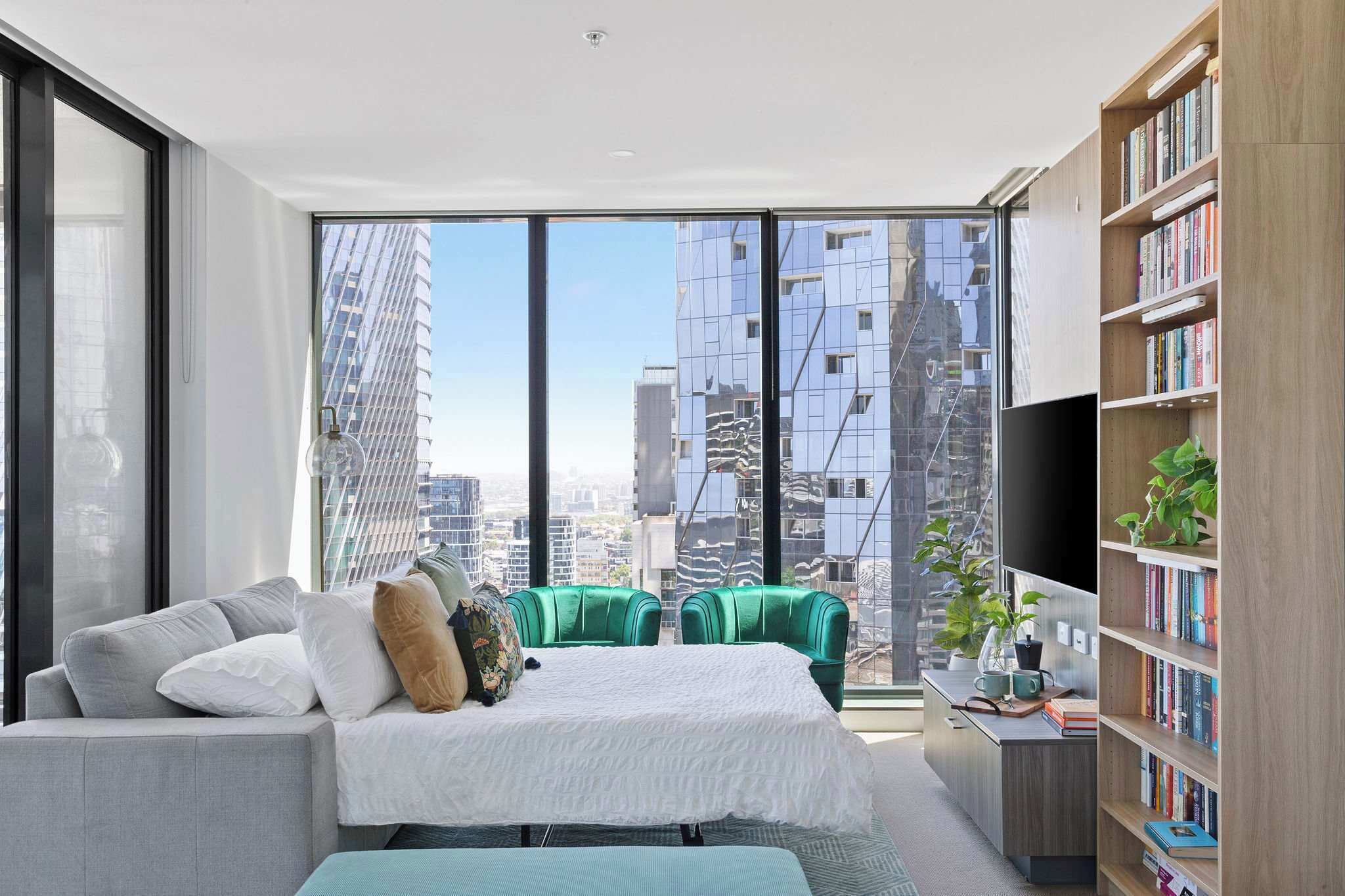 Une chambre moderne avec une grande fenêtre, un lit, un canapé vert, une bibliothèque et un bureau agrémenté d’une plante.