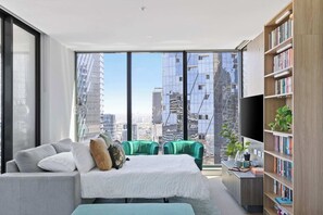 Une chambre moderne avec une grande fenêtre, un lit, un canapé vert, une bibliothèque et un bureau agrémenté d’une plante.