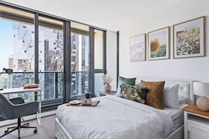 A modern bedroom with a large window, a bed with white bedding, a desk with a chair, and artwork on the wall.