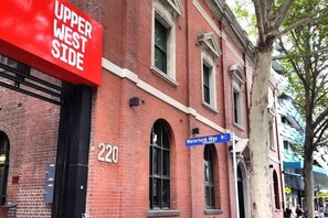Un bâtiment en briques rouges sur lequel on peut lire l’inscription « Upper West Side », ainsi qu’un panneau de signalisation bleu indiquant « Watertank Way ».