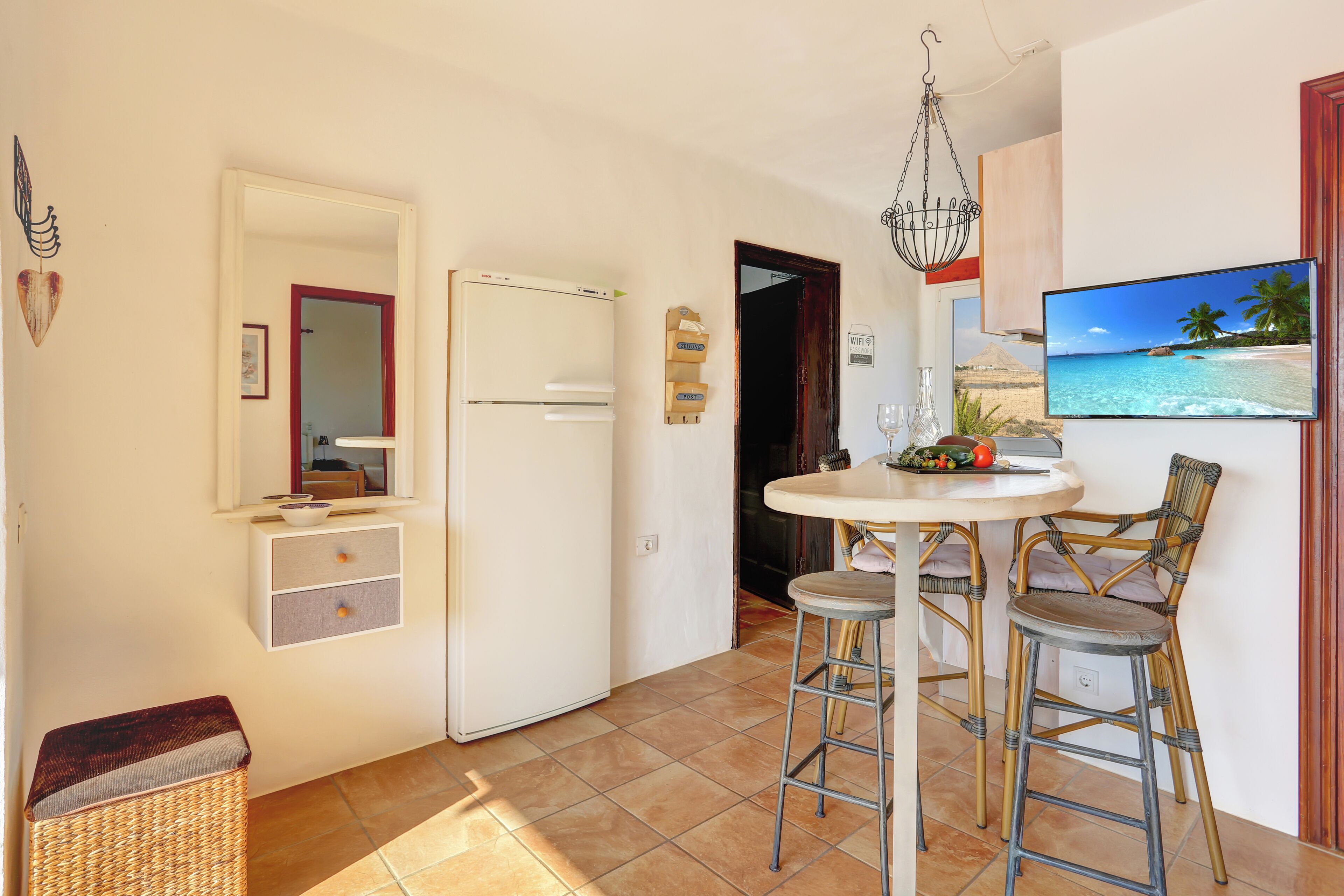 Cocina compacta con refrigerador, una mesa de comedor con dos sillas, lámpara colgante y un televisor de pared mostrando una escena de playa.