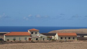 Il s’agit d’une propriété située sur la côte, comprenant trois bâtiments, chacun coiffé d’un toit recouvert de tuiles rouges. Elle est construite sur un terrain aride et rocheux et offre une vue sur la mer en arrière-plan.