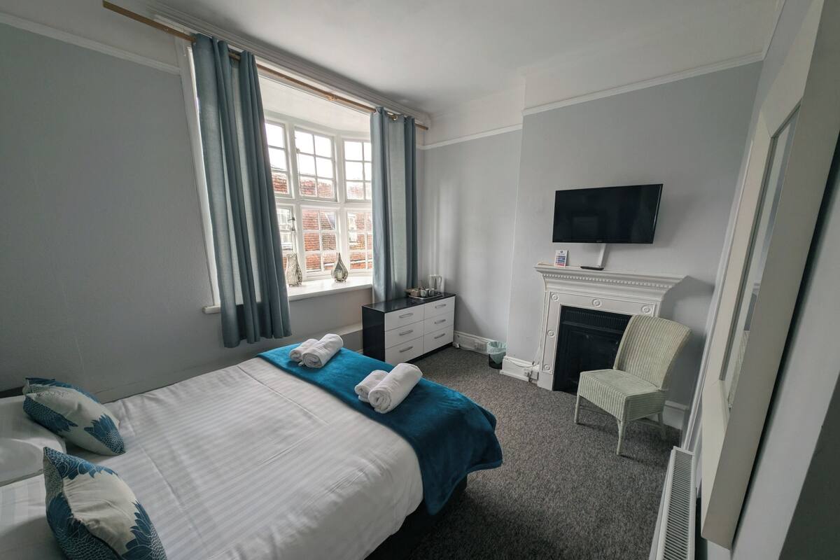 Una habitación de hotel con cama, tocador, televisor, chimenea y ventana con cortinas.