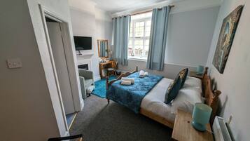 Un dormitorio con cama, escritorio, silla, televisión, ventana con cortinas y radiador.