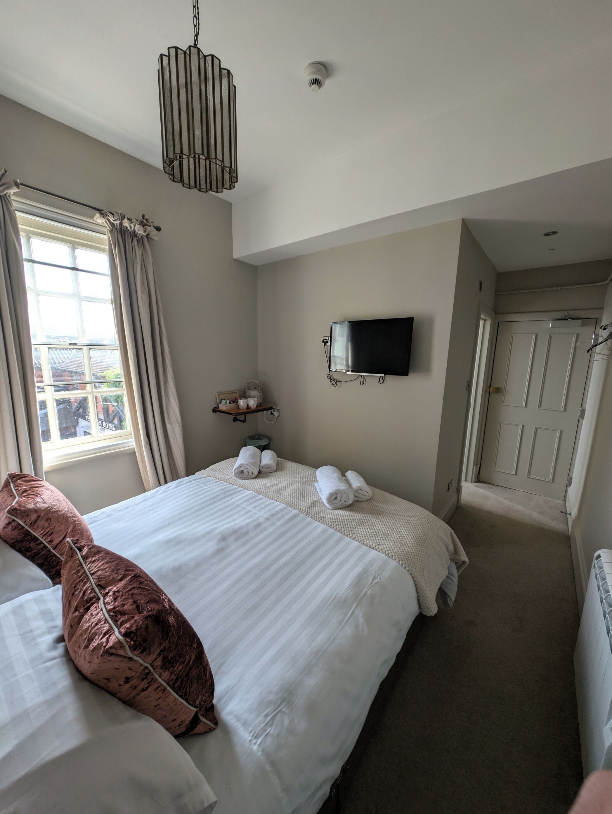 A hotel room with a large bed, a television mounted on the wall, a window with curtains, and a door leading to another room.