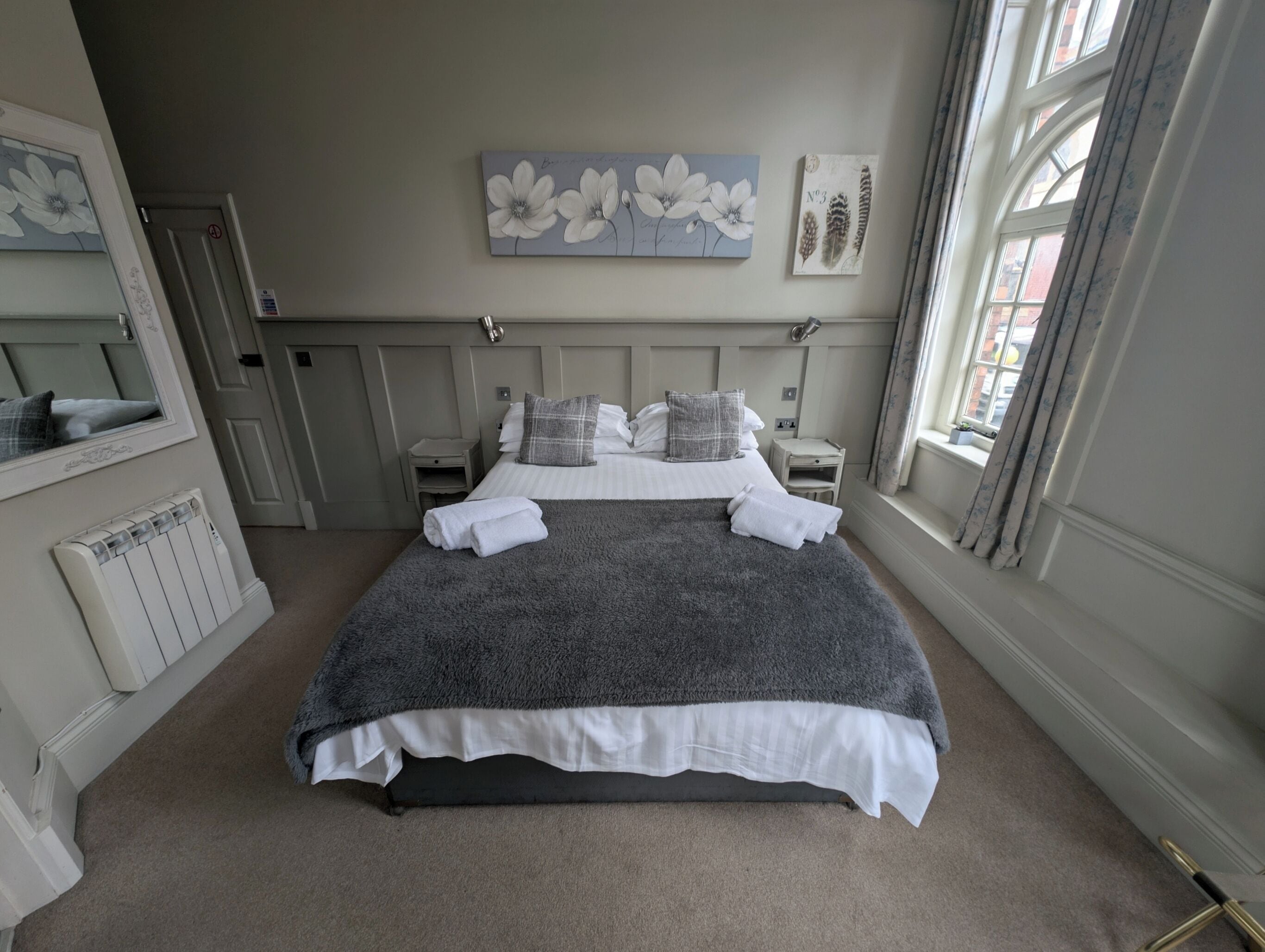 Una cama bien hecha con una manta gris, almohadas blancas y un plaid gris. Sobre la cama, hay un cuadro de flores en la pared, y a la derecha, una ventana con cortinas.