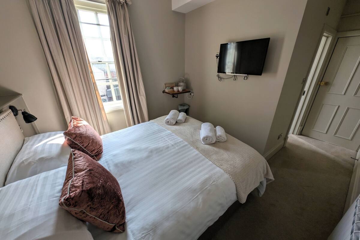 Habitación de hotel con dos camas, un televisor de pantalla plana montado en la pared, una ventana con cortinas y un estante pequeño con artículos de aseo.