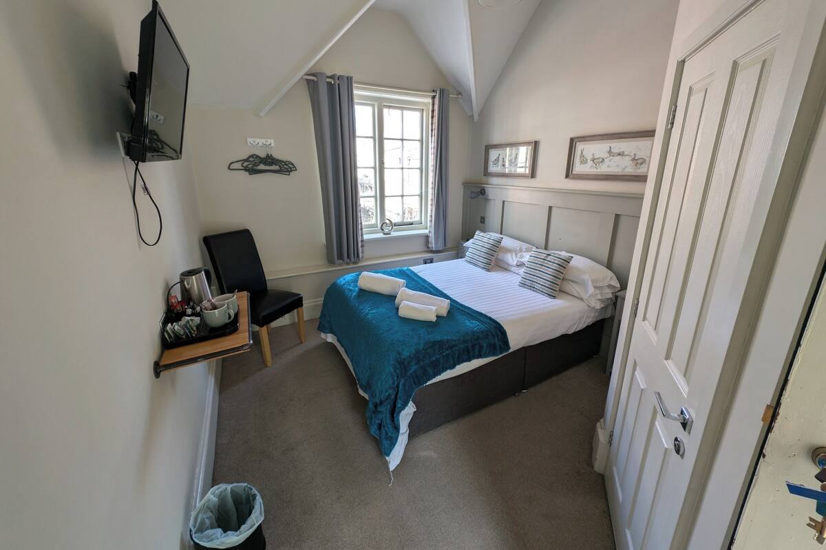Un dormitorio con cama, una silla, televisión, ventana con cortinas y puerta.