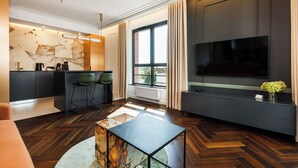 Ein modernes Wohnzimmer mit Fischgrätparkett, einem Marmortisch, einer Couch, einem Fernseher auf einer dunklen Konsole und einer Küchenzeile mit Steinrückwand und Barhocker.