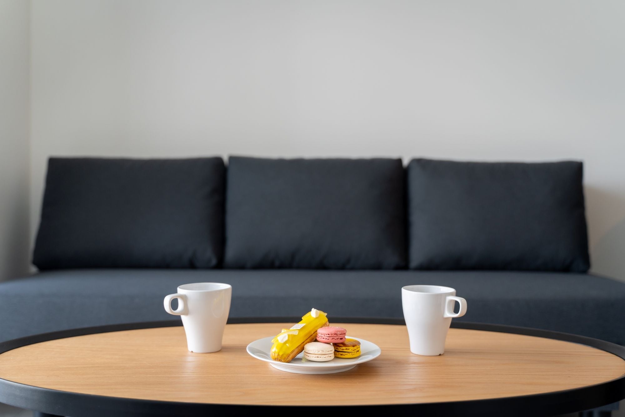 A round wooden table with two white coffee cups and a plate of macarons in front of a dark grey sofa with three cushions.