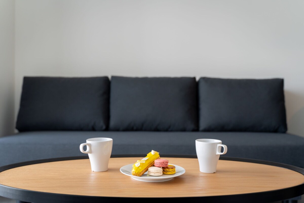 A round wooden table with two white coffee cups and a plate of macarons in front of a dark grey sofa with three cushions.