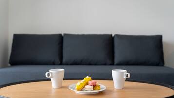 A round wooden table with two white coffee cups and a plate of macarons in front of a dark grey sofa with three cushions.