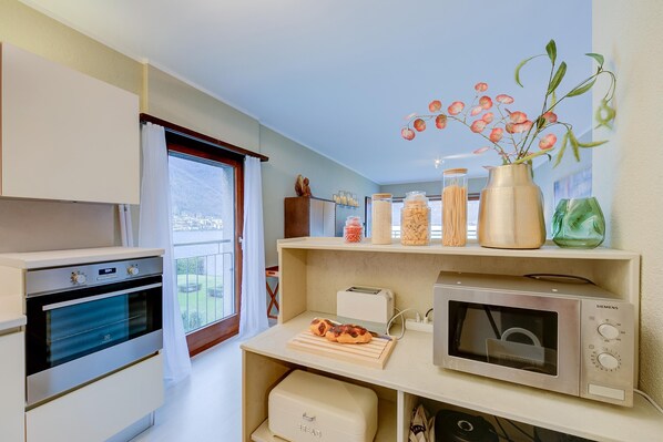 A compact kitchen with an oven, microwave, and a small countertop with a cutting board and a toaster. - Emotion Lake View in Capo Lago (Mendrisio)