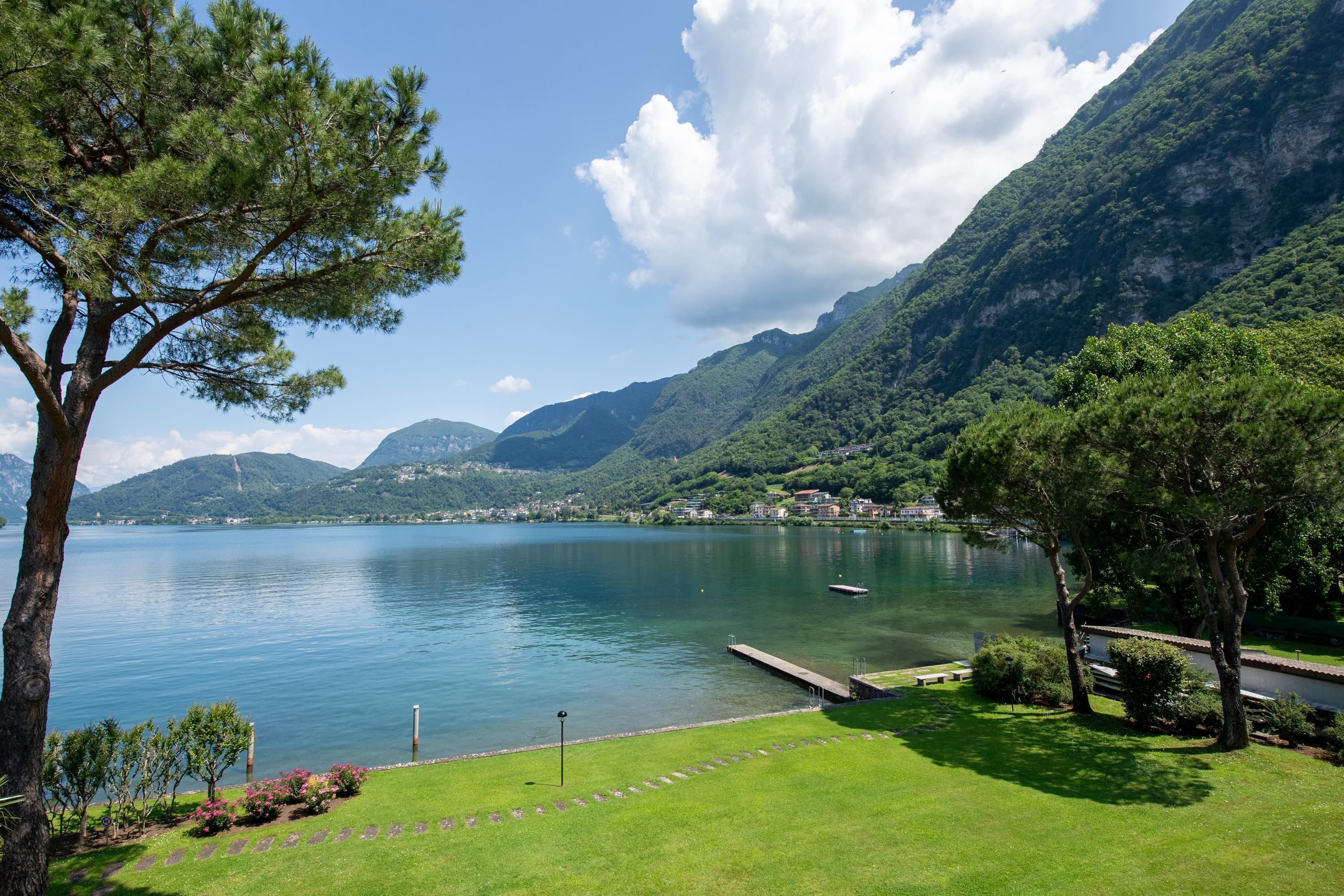 A serene lakeside view with a dock, lush greenery, and mountains in the background.
