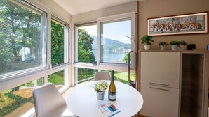 Un coin repas avec une table dressée pour deux personnes, une bouteille de vin et un vase de fleurs. De grandes fenêtres offrent une vue sur un lac et la verdure environnante.