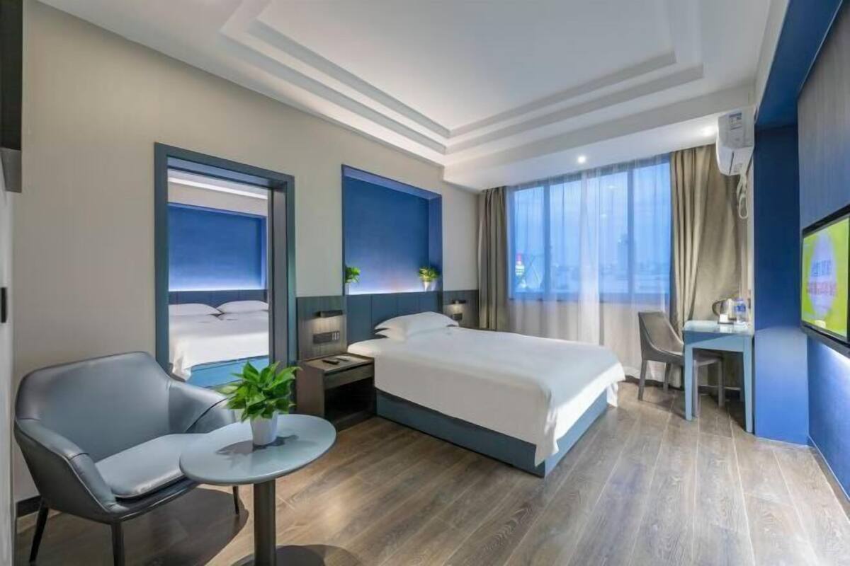 Ein modernes Hotelzimmer mit Bett, Schreibtisch, Stuhl, einem kleinen Tisch mit Pflanze und Blick auf die Stadt durch große Fenster.